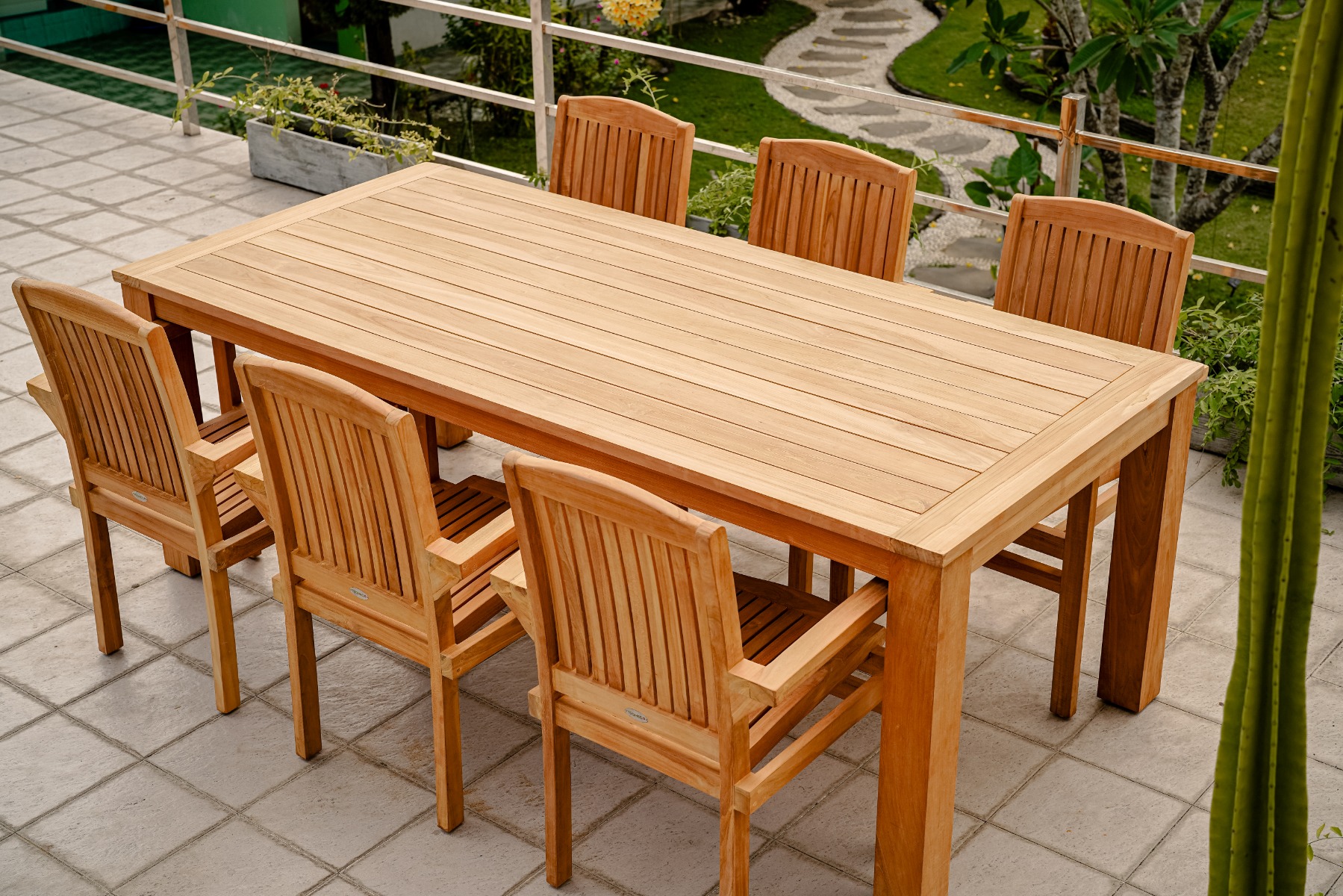 Een sfeervolle foto van een grote, rechthoekige teakhouten tuintafel met zes bijpassende teakhouten stoelen op een zonnig, betegeld terras. De tafel is stijlvol gedekt met linnen lopers, servies, houten schalen met fruit en brood, en kruidenplanten. De st - alternate view